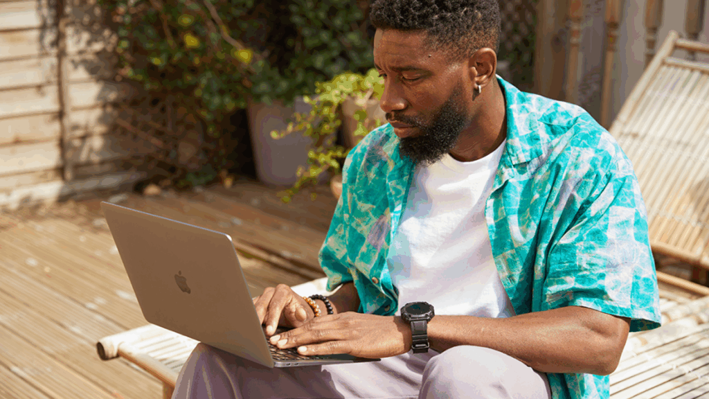A man in a patterned shirt is working on a laptop outdoors, searching for how to start a print-on-demand business.